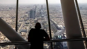A man is looking out of a large window in a tall building a a sprawling cityscape below.