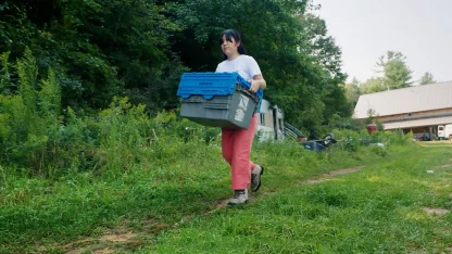 Student, Cassia Park, carries a stack of tote boxes down a path away from a barn.
