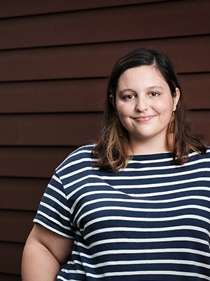 Claire Contreras in a black-and-white striped top, smiling in front of a dark red siding background