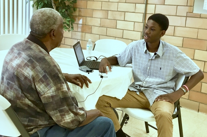 A young Black man sits at a computer and listens to an older Black man.