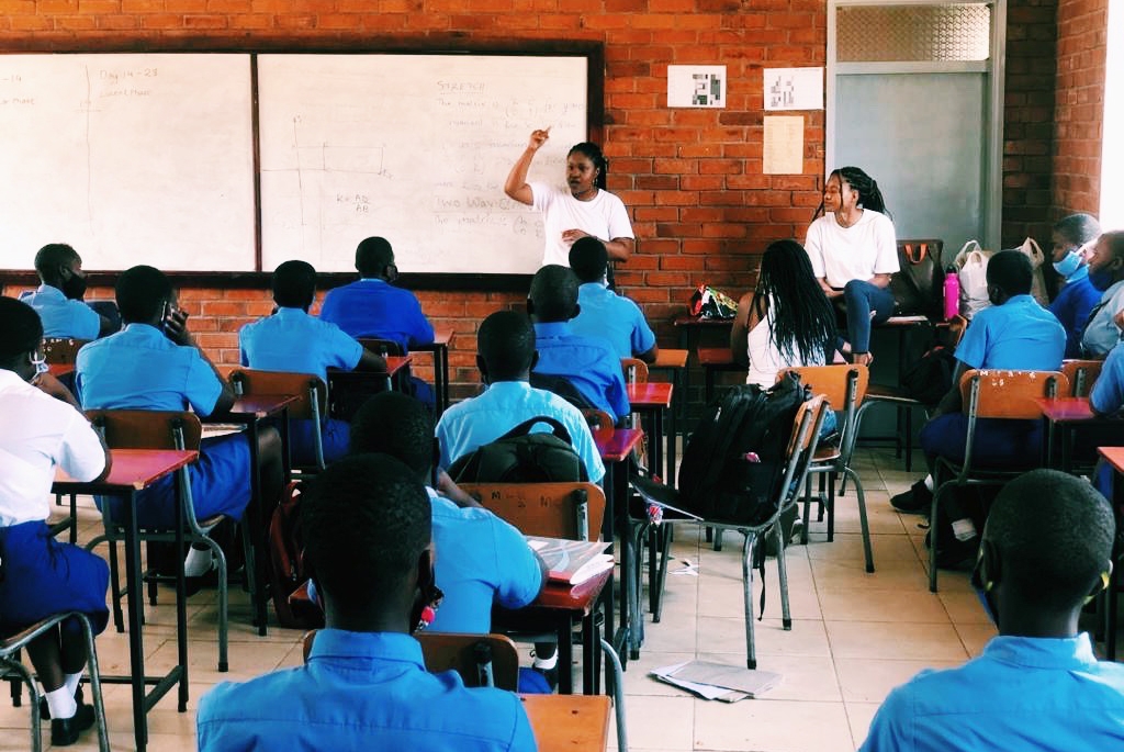 Marie-Simone Kadurira teaching at a whiteboard at the front of a full classroom.