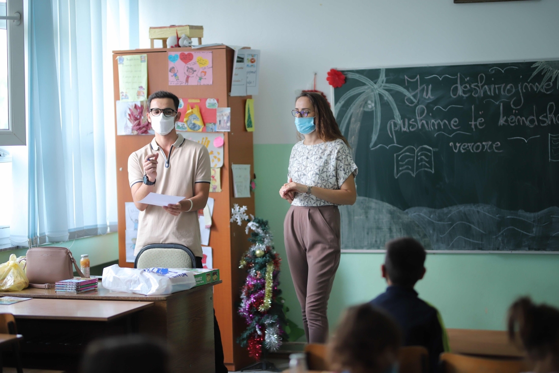Two people in business casual dress and surgical masks stand at the front of a classroom and address a group of young students