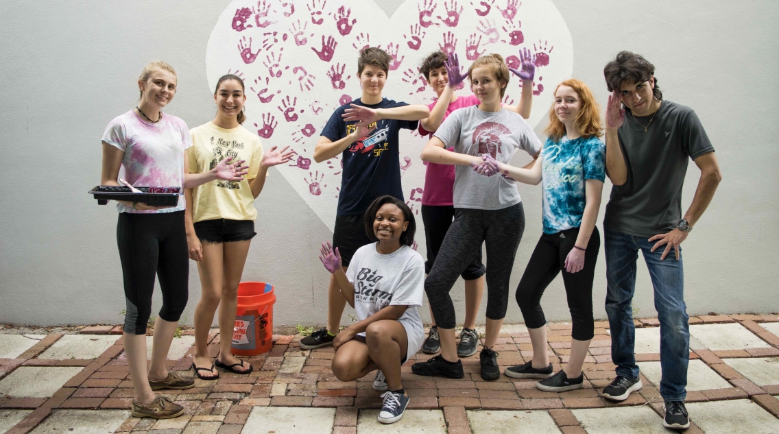 A group of teenagers of various skin tones stand in front of a heart-shaped mural they completed