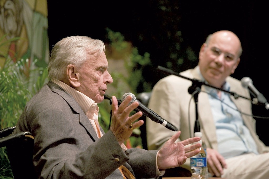 Wearing a brown blazer and orange shirt, Gore Vidal speaks into a microphone on stage sitting next to Jay Parini, who wears a tan suit and blue shirt.