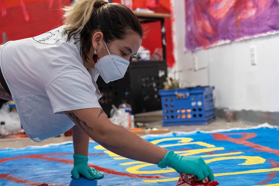 Wearing a mask, white shirt, and blue gloves, Sienna Bucu paints on a large mural of blue, yellow, and red.