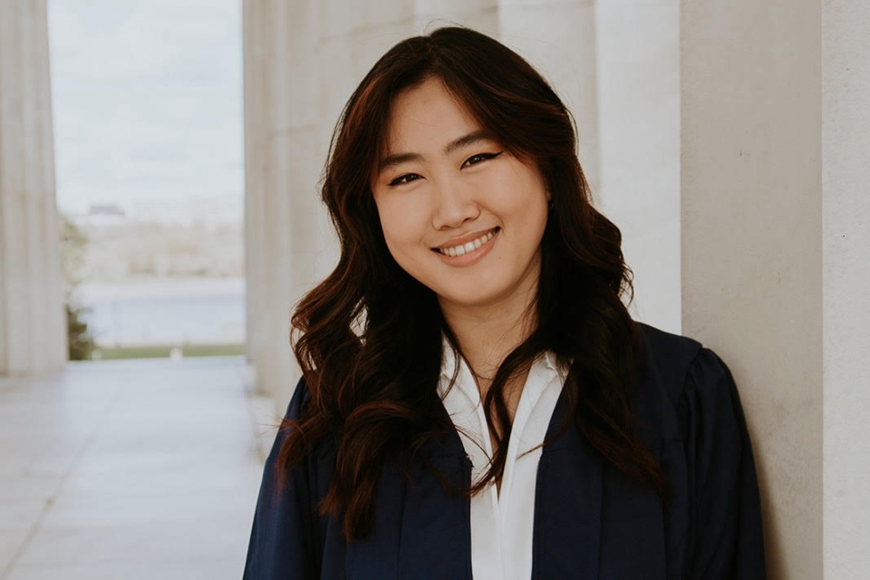 Emily Zhang, wearing a navy blazer and white blouse, smiles for the camera in front of a series of marble columns opening up to the waterfront.