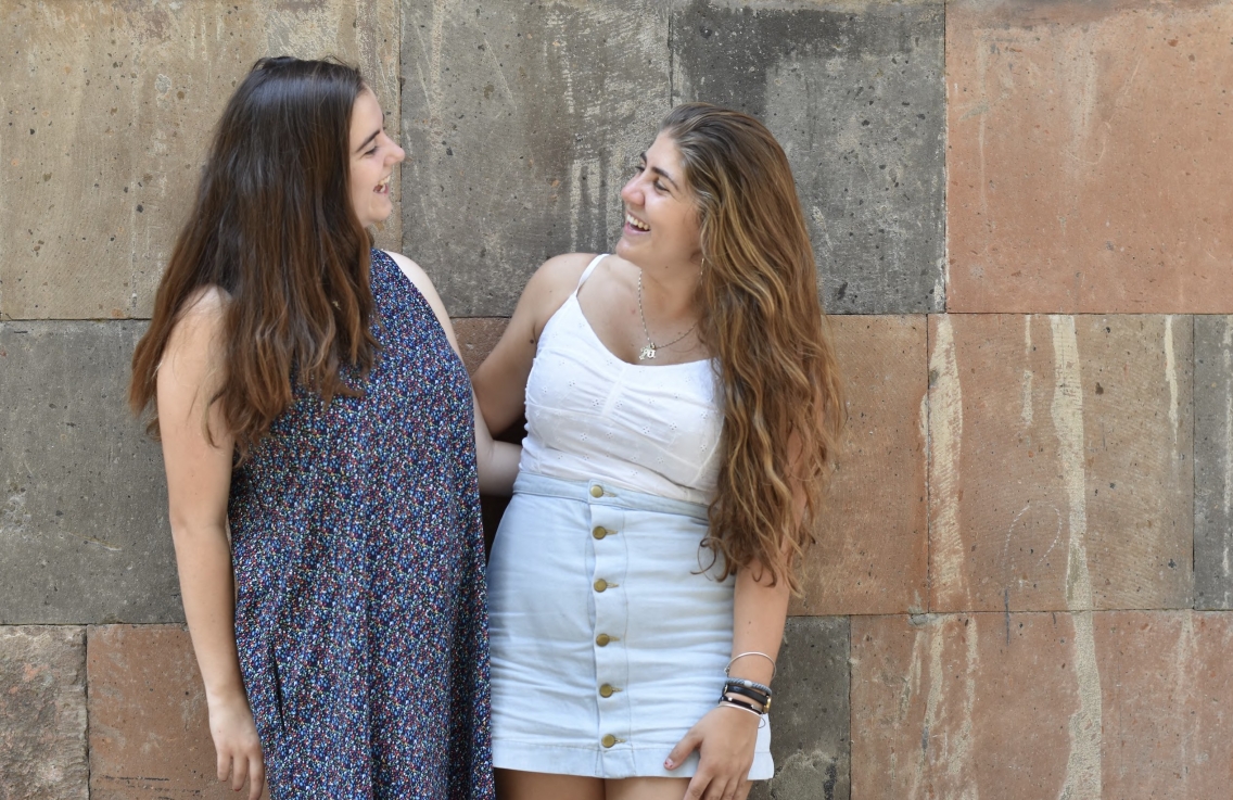 Ani Schug and Anoush Baghdaserrian look and smile at each other with their arms linked, standing in front of a rock wall.