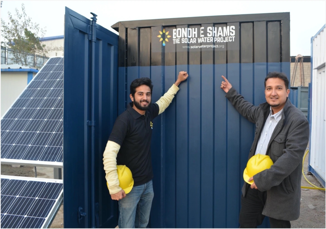 Two men in dark clothing holding yellow construction hats stand in front of and point to a blue and black 'Oasis Box' meant to clean water.