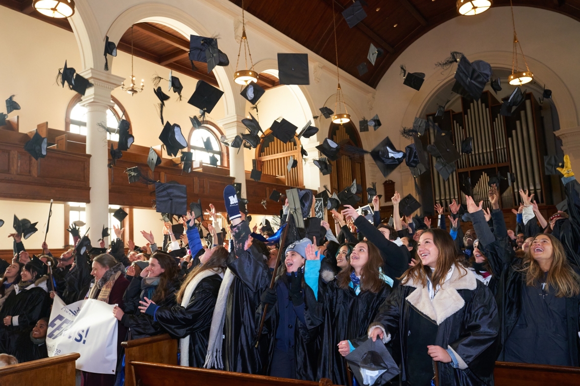 Members of the Class of 2022.5 inside the brown and white Middlebury Chapel toss their black graduation caps in the air 