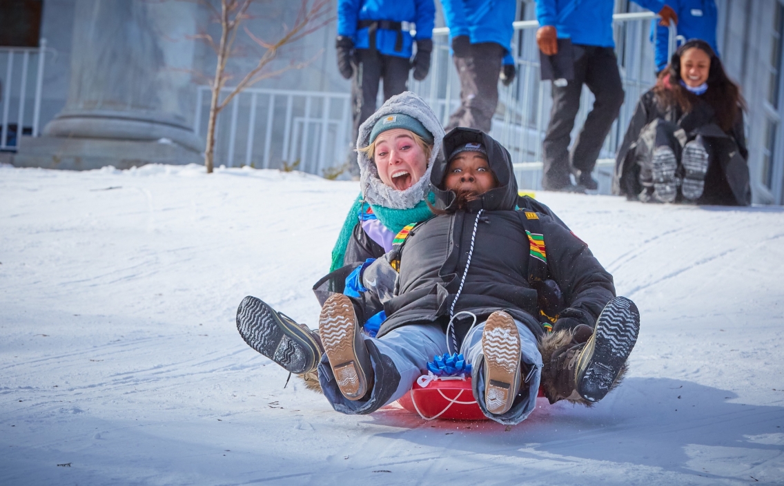 Two people in winter jackets and hats scream with delight as they ride a red toboggan down a snowy hill.