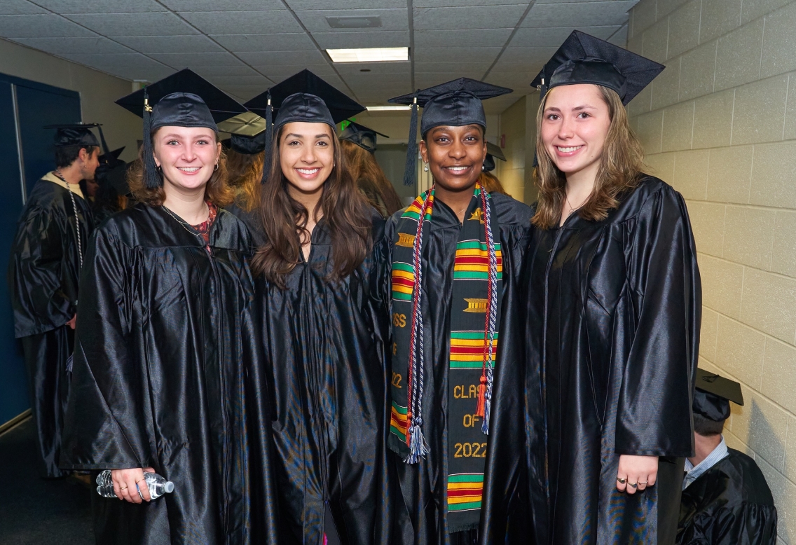 Four graduates, wearing black caps and gowns, pose together in a hallway.