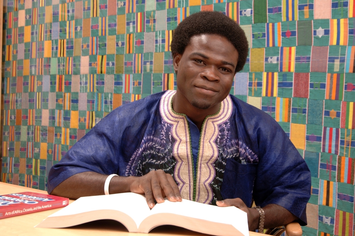 Joseph Kaifala, wearing a cream and blue colored shirt, poses sitting at a desk with his hand on a book, flanked by a multi-colored wall.