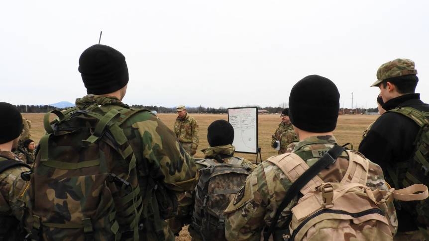 ROTC students stading with their backs to the camera, in full camp gear and rucks, listening to an instructor standing in a field with a dry-erase board.