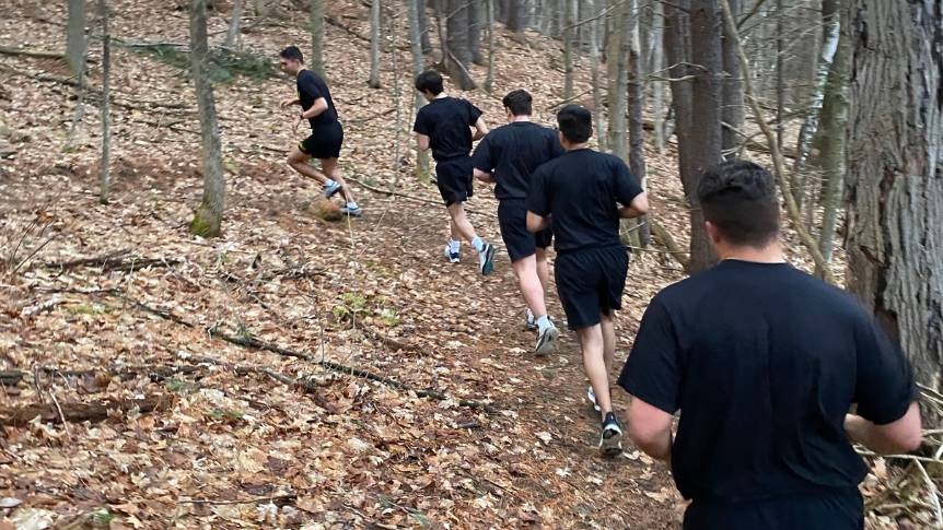 5 ROTC students doing a training run in the woods in the fall. They are all wearing black shorts and black t-shirts.