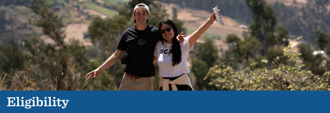 A young man in a black shirt and a young woman in a white shirt walk up a hill with their arms around each other, waving to the camera. Below the picture a blue graphic footer features the word "Eligibility" in white.