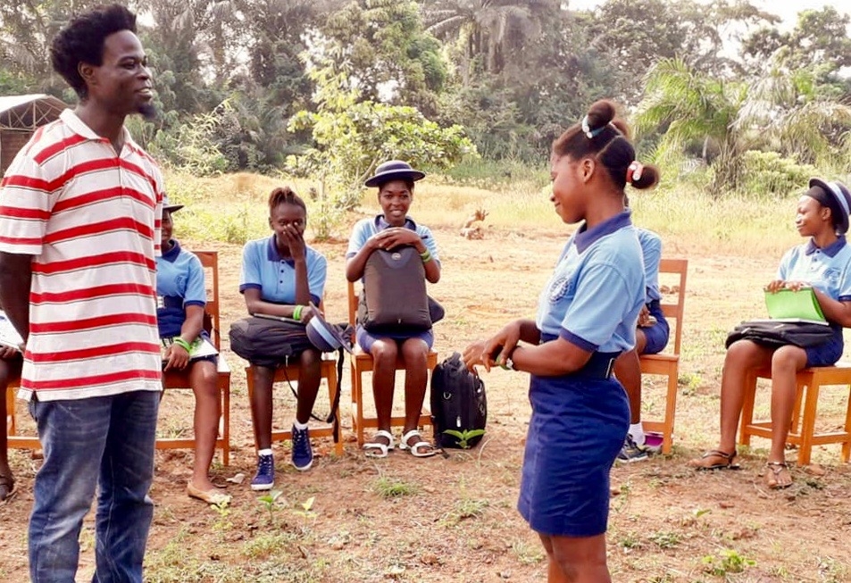 Joseph Kaifala, wearing a red and white striped shirt and jeans, stands in the middle of a seated circle of female students with a female student dressed in a blue uniform