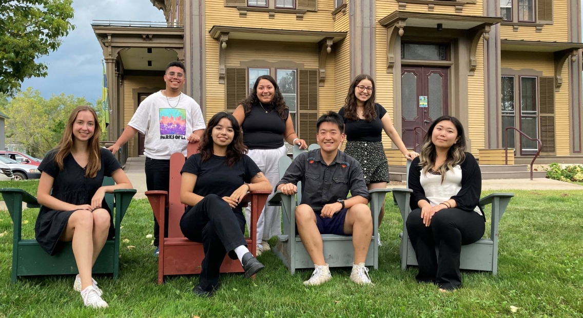 A group of seven smiling students posing for a groupo photo. 4 are sitting in adirondack chairs and 3 are standing behind them. They are in front of Kitchel House, the home of the Center for Careers and Internships.