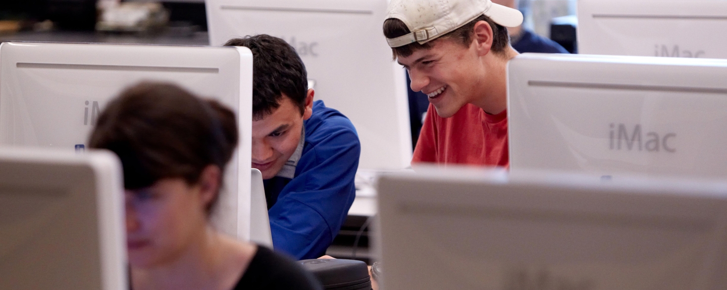 People working in a computer lab of iMacs. One woman is in a front row and behind her is a row with two men looking at a laptop.