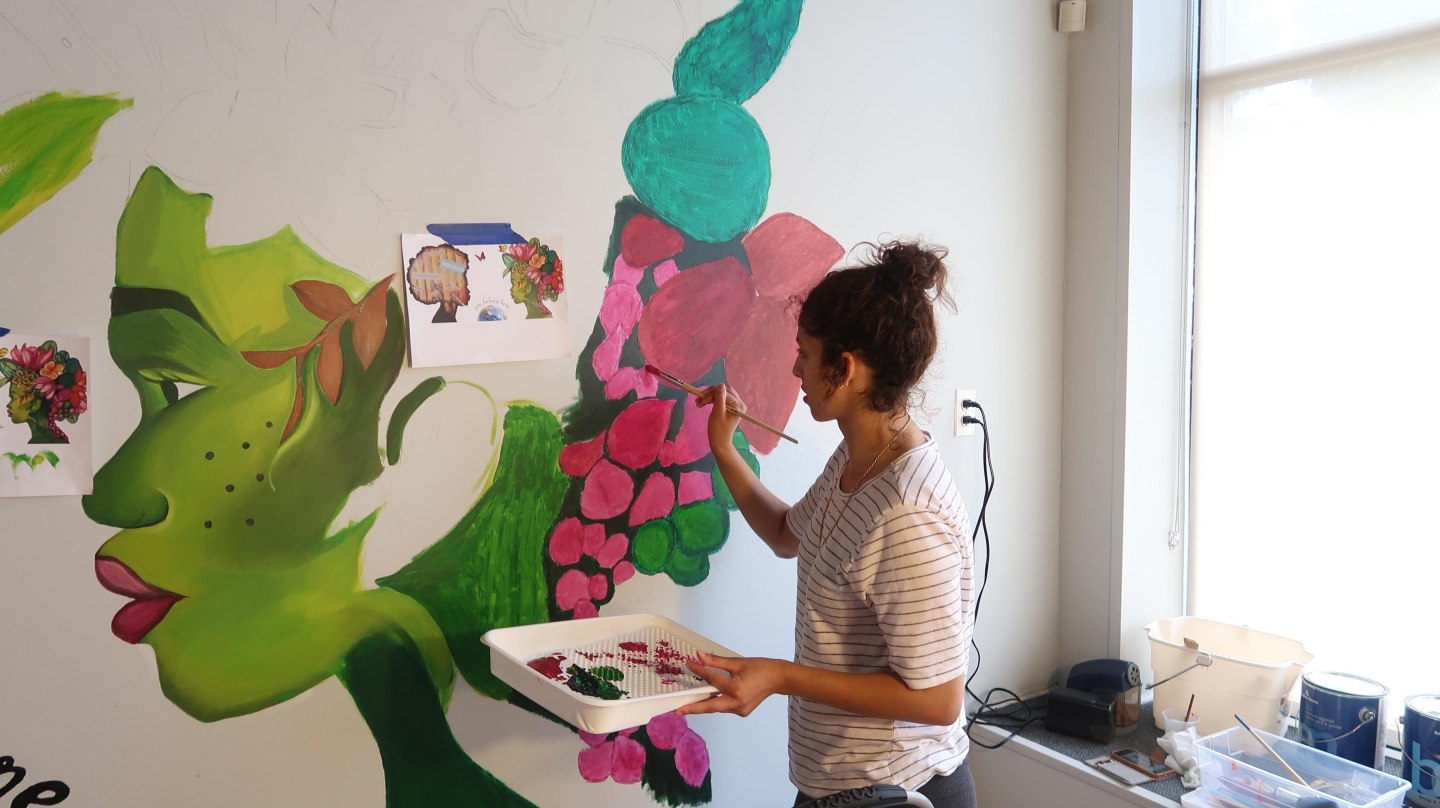 A woman wearing a white shirt with her hair pulled in a bun works with a small paintbrush on a mural of a green-skinned woman with multi-colored flowers instead of hair.