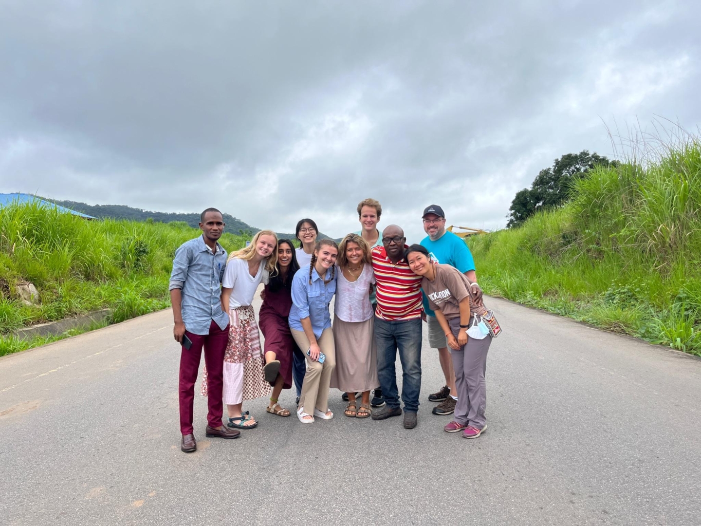 A group of people of various heights, skin tones, and ages pose with their arms around each other in the middle of a paved road that cuts through lush green hills.