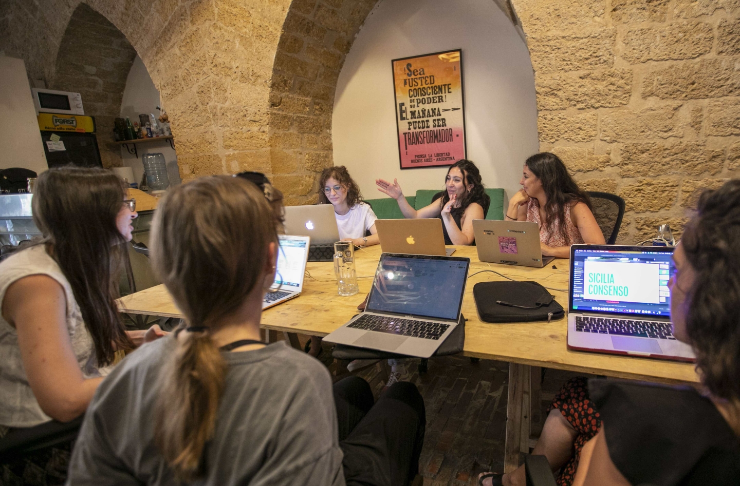 A group of women of various ages sit at a conference table with their laptops in the midst of an animated discussion.