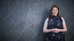 Wearing a blue floral dress and crossing her arms across her stomach, Hannah Harris poses for a portrait in front of a tiled, dark gray, wall.