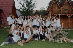 A group of young people, all wearing the same white t-shirt with a green and blue emblem, pose for a silly group photo in front of a log cabin.