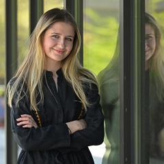 Blonde-haired Olha Vasyliv, wearing a black jacket and crossing her arms, poses for a portrait in front of a reflective glass wall.