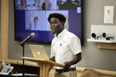 Joseph Kaifala, wearing a short-sleeve white button-down shirt and khaki pants, stands and speaks at a wooden podium topped by a laptop.