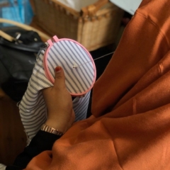 Facing away from the camera, a women in an orange headscarf pulls a needle and thread through a new cross-stitch project.