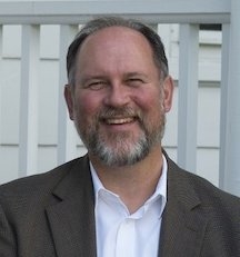 smiling white man with beard wearing white collared shirt and brown suit jacket