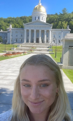 A young woman with blond hair is smiling and taking a selfie in front of the Vermont Statehouse. It is summer and the sky is blue.