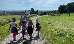 Photo of a group of children headed down a dirt path with the Shelburne Farms vista in front of them. it is summer, the children are holding hands, and they are led by a Middlebury student. They are headed toward the barn.