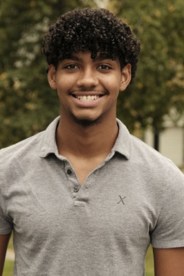 A young man of color with curly brown hair, and a brown polo shirt smiles into the camera
