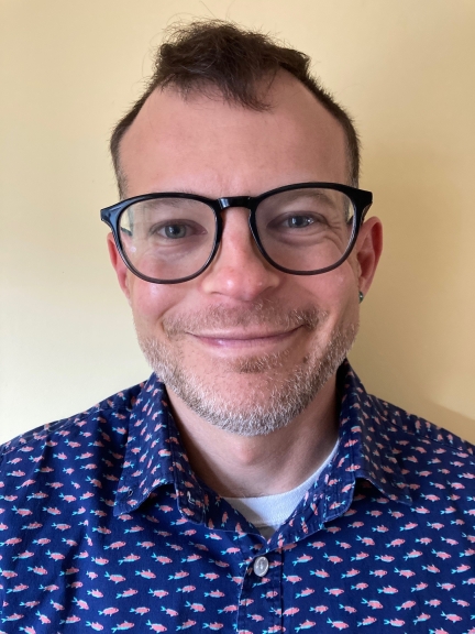 Headshot of Nick wearing black-rimmed glasses, a blue shirt and a smile against a beige backgrond