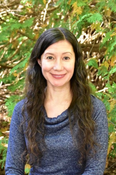 A woman with long, dark wavy hair smiling in front of a natural background.
