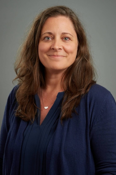 A white woman with long brown hair, blue eyes, and a blue blouse and cardigan stands in front of a grey background.