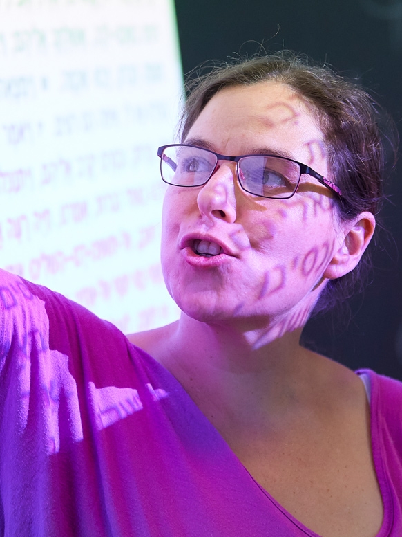 A women looks to her right as Hebrew text is projected over her face and on a screen.