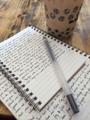 handwritten paper journal with spiral binding; pen lying on top with coffee cup nearby