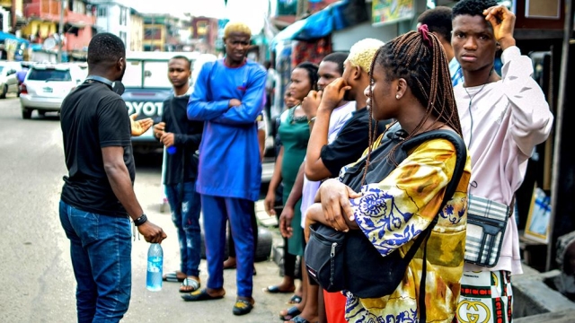 Wearing a yellow shirt and cradling a black backpack, Amara Ugochukwu stands in front of a group of people mingling in front of an outdoor market.