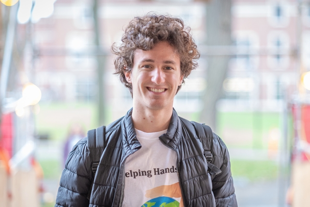 Jonathan Tamen poses smiling outside wearing a slate grey puffer jacket and white t-shirt with the text "helping hands" in black just visible.