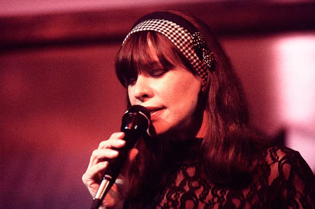 Astrud Gilberto performing in the mid 1990s; she holds a microphone close to her face and has a wide patterned headband over her hair.