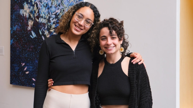Valeska Kohan and Amanda de Souza, both wearing black shirts, pose smiling for the camera with their arms around each other in front of a white wall and multi-hue blue painting.