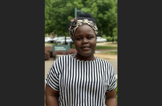 Rabecca Ndhlovu, wearing a black and white striped shirt and a brown headscarf, poses for a photo in front of a tree-filled background.