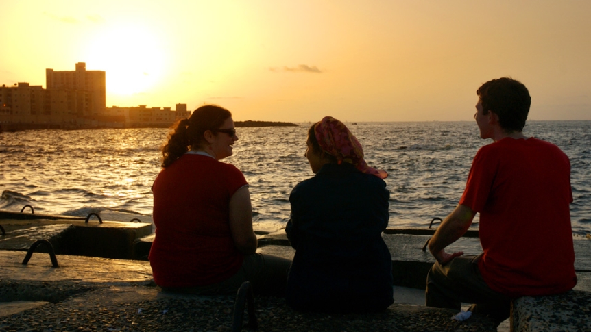 Three students sitting on breakwall near ocean at sunset.