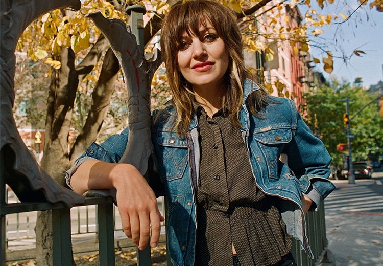 Portrait of singer Anais Mitchell. She is leaning against a metal fence beneath a tree.