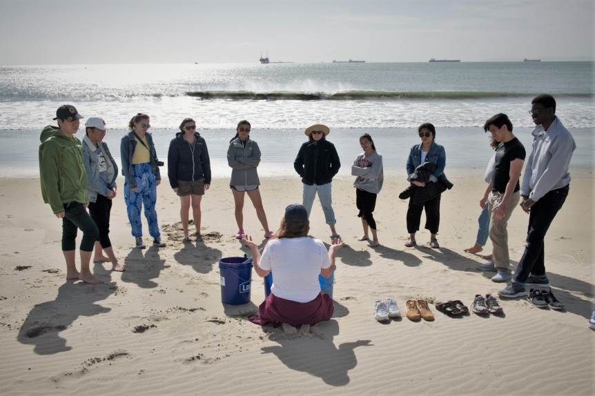 A group of students stand barefoot on the beach, huddled around a MAlt leader.