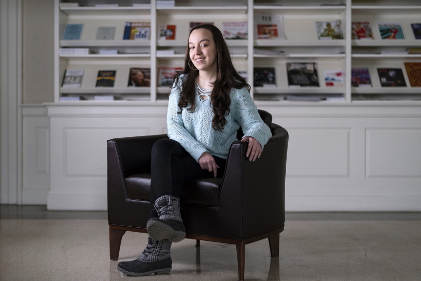 Giovanna Novi, wearing a light blue sweater, black pants, and gray boots, smiles and sits in a brown chair placed in front of white bookshelves.