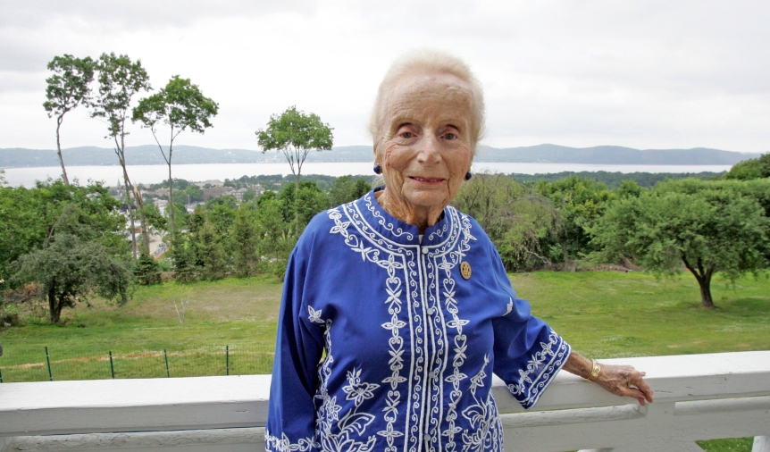 Wearing a blue and white blouse, Kathryn W. Davis smiles fro the camera while leaning on the white railing of a porch overlooking a green lawn and forest.