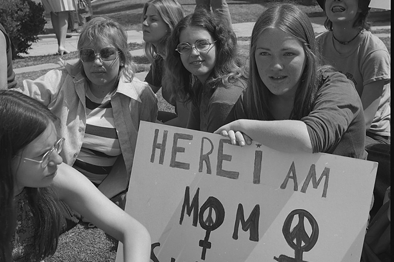 Black and white photo of four young women seated around a protest sign, which reads "Here I am Mom." There are two symbols side-by-side that incorporate the symbol for Venus/women and a peace sign.